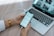 A person is holding a smartphone displaying a to-do list or task management app near an open laptop. The phone is connected to the laptop with a cable. The laptop is on a wooden desk showing web pages on the screen. The person is wearing a white sleeve and a gold watch.
