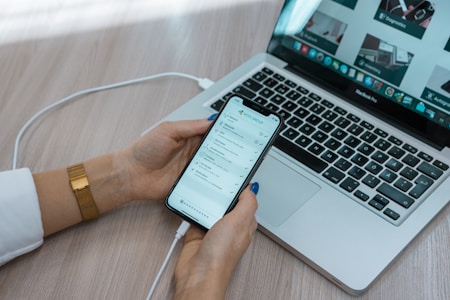 A person is holding a smartphone displaying a to-do list or task management app near an open laptop. The phone is connected to the laptop with a cable. The laptop is on a wooden desk showing web pages on the screen. The person is wearing a white sleeve and a gold watch.