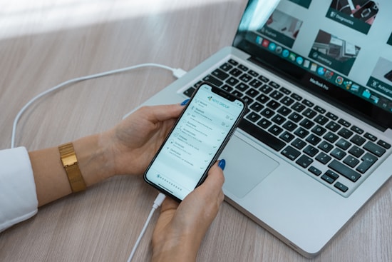 A person is holding a smartphone displaying a to-do list or task management app near an open laptop. The phone is connected to the laptop with a cable. The laptop is on a wooden desk showing web pages on the screen. The person is wearing a white sleeve and a gold watch.