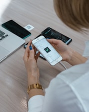 A person with blue nail polish is holding a smartphone displaying a checklist with a green checkmark. The right wrist features a gold watch. A laptop, another smartphone, and a QR code on a piece of paper are visible on the wooden table top.