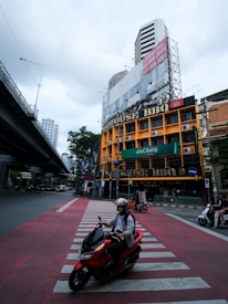 An urban scene featuring a multi-story building with large signage indicating a BBQ restaurant. The building is situated at a major intersection with visible zebra crossings. Several motorcyclists are riding across the street, and an overhead highway runs parallel to the scene. The sky is overcast, adding a muted tone to the image.