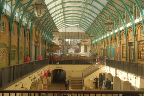 An interior view of a spacious marketplace with a high arched glass ceiling supported by teal metal beams. The building features brick walls with decorative details and large hanging lanterns. Several shops are visible along the sides, with people walking and standing on the upper level. The lower level has more shops and several people are seen moving about or stopping near a staircase.