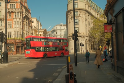 A classic red double-decker bus is turning at an intersection in a city with historic architecture. The buildings feature detailed facades with brick and stone elements. Several pedestrians are walking on the sidewalk, including a person wearing a trench coat. The street is relatively calm, and there is a sign for a restaurant on the right.