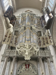 A large and ornate pipe organ is mounted within an interior space that features classical architecture. The organ is embellished with intricate designs and gilded accents. Surrounding the organ are decorative banners and columns, adding to the grandiose and historical ambiance.