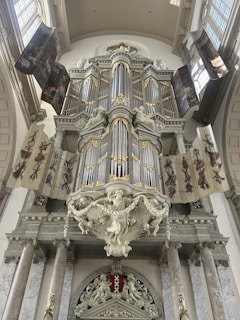 A large and ornate pipe organ is mounted within an interior space that features classical architecture. The organ is embellished with intricate designs and gilded accents. Surrounding the organ are decorative banners and columns, adding to the grandiose and historical ambiance.
