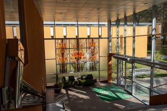 A spacious interior area features a large stained glass window with colorful geometric patterns in red, orange, and yellow hues. Two modern green chairs are positioned on a brown tiled floor near a potted plant. The room is bright due to ample natural light streaming in through tall glass walls, which also offer a view of the greenery outside. A green mat is placed at the entrance, and the architectural design includes a high ceiling and a metallic staircase visible on the left.