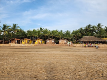 A beach scene with several huts and small buildings lined at the edge. Palm trees are visible behind the structures, creating a tropical setting. The sand stretches across the foreground, and some colorful umbrellas and chairs can be seen near the huts.