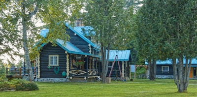 A charming log cabin is nestled among tall trees. The cabin features dark wood logs with contrasting light-rounded ends. The roof is a bright blue metal, complementing the serene natural setting. A swing is visible in the front yard, and the grassy area is well-maintained with patches of shadow from the surrounding trees. A small porch with flowers adds a touch of color to the serene environment.