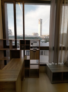 An interior scene featuring a wooden shelving unit with various compartments. A glass pitcher is placed on one of the compartments. The shelving unit is positioned in front of a large window with sheer curtains pulled to the side. Outside the window, a tall clock tower and several buildings with white walls and green roofs are visible under a partly cloudy sky.