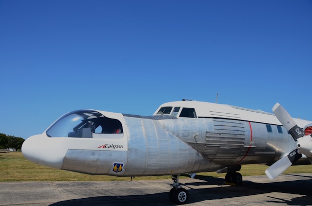 A silver aircraft with a white nose is parked on an airfield under a clear blue sky. The aircraft has a streamlined design, visible cockpit, and labeled with 'Calspan'. The surrounding area includes grassy fields and paved surfaces.