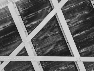 An angled view of an industrial structure featuring a series of diagonal and horizontal metal beams bolted together, set against a backdrop of weathered wooden panels.