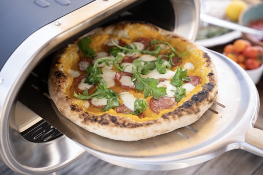A freshly baked pizza with melted cheese, pepperoni slices, and fresh arugula is being taken out of a pizza oven on a metal pizza peel. The crust is golden with slightly charred edges, and the colorful toppings stand out against the light background.