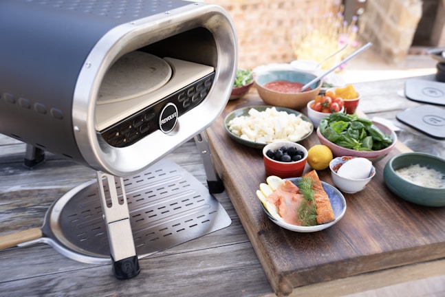 A portable pizza oven is placed on a wooden surface, surrounded by various bowls containing ingredients such as tomatoes, arugula, olives, cheese, and salmon. There is also a metal pizza peel leaning against the pizza oven. The setting appears to be outdoors with a blurry background of bricks and plants.