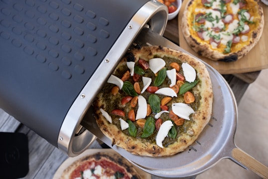 A freshly baked pizza covered with sliced mozzarella, cherry tomatoes, and basil leaves on a silver peel is being taken out of a pizza oven. There are other pizzas in the background on wooden boards, topped with various ingredients.