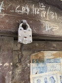 A close-up view of a metal padlock attached to a loop on a wooden door. The padlock has visible text and appears worn, indicating age. Chalk writings are on the upper part of the door, and faded posters featuring text and images are pasted below.