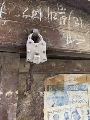 A close-up view of a metal padlock attached to a loop on a wooden door. The padlock has visible text and appears worn, indicating age. Chalk writings are on the upper part of the door, and faded posters featuring text and images are pasted below.