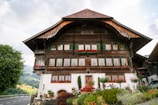 A traditional wooden Swiss chalet with a steeply pitched roof and numerous windows adorned with green shutters. The exterior features wooden beams and floral arrangements, particularly red geraniums, beneath the windows. The chalet is surrounded by a lush garden with various colorful flowers, situated next to a road with glimpses of a scenic landscape in the background, including hills and trees.