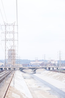 A concrete-lined river channel with a narrow flow of water runs through an urban landscape. Industrial structures, bridges, and power lines are visible alongside the channel. The backdrop includes buildings and hills under an overcast sky.