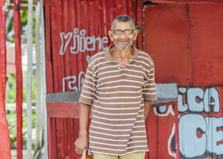An elderly man with a gentle smile stands in front of a red corrugated metal structure. He is wearing a brown and white striped polo shirt and glasses. The background features red and white painted designs with some unreadable text.