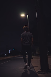 A person walking alone on a dimly lit street at night under a streetlamp. The street is mostly empty, and the surroundings are dark, creating a sense of solitude.