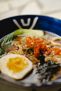 A close-up view of a bowl of ramen featuring broth, noodles, sliced red chili peppers, a halved soft-boiled egg, green vegetables, and a piece of seaweed. The dish is served in a blue bowl with decorative patterns.