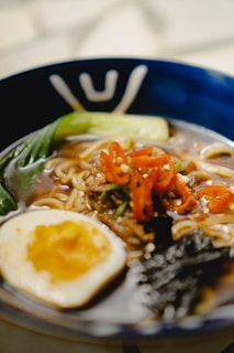 A close-up view of a bowl of ramen featuring broth, noodles, sliced red chili peppers, a halved soft-boiled egg, green vegetables, and a piece of seaweed. The dish is served in a blue bowl with decorative patterns.
