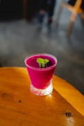 A vibrant magenta-colored smoothie served in a textured glass topped with a small sprig of fresh mint. It rests on a warm-toned wooden surface with soft, natural lighting creating a cozy atmosphere.