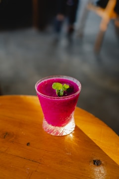 A vibrant magenta-colored smoothie served in a textured glass topped with a small sprig of fresh mint. It rests on a warm-toned wooden surface with soft, natural lighting creating a cozy atmosphere.