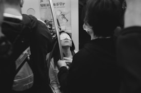 A woman with a sleep mask over her eyes is sitting on public transportation, surrounded by other passengers standing nearby. The environment is crowded, and the passengers appear to be either standing or holding onto poles.