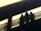 Three indistinct shadows cast on a surface, appearing to stand next to a railing. The lighting creates a stark contrast, with bright areas highlighting the shapes.