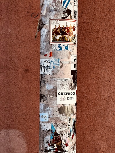 A vertical pole is covered with various stickers and posters, including promotional images and text in different languages. The background wall is of a reddish-brown color.