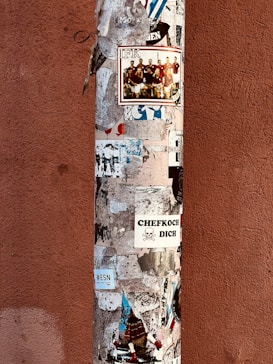 A vertical pole is covered with various stickers and posters, including promotional images and text in different languages. The background wall is of a reddish-brown color.