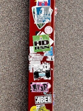 A red surface is covered with multiple colorful stickers, including logos, text, and designs. The background is a textured gray surface.