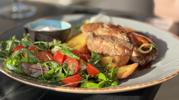 A plate of food featuring a well-cooked piece of meat, accompanied by roasted potatoes and a fresh green salad with cherry tomatoes. A small bowl, possibly containing a sauce or dip, is also present on the plate.