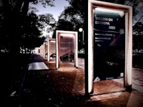 A series of illuminated frames line a pathway, each containing posters with text and images. The scene is set in a park-like area with trees casting shadows in the background. The lighting gives a dramatic and somewhat mysterious atmosphere.