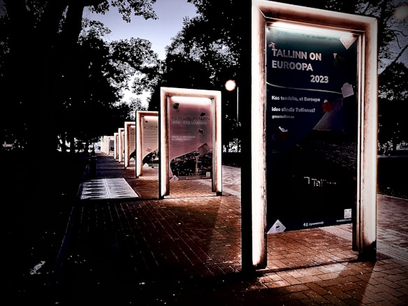 A series of illuminated frames line a pathway, each containing posters with text and images. The scene is set in a park-like area with trees casting shadows in the background. The lighting gives a dramatic and somewhat mysterious atmosphere.