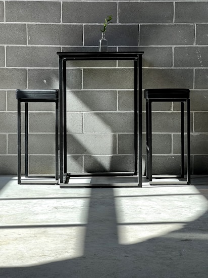 A minimalist setup featuring a small metal table with two matching stools on a concrete floor. A single branch in a glass vase sits atop the table, casting a distinct shadow on the wall made of gray bricks. The sunlight streaming in creates geometric patterns of light and shadow across the floor and wall.