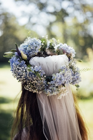 A floral crown made of light blue hydrangeas, white baby's breath, and greenery atop long, flowing hair. The image is taken from behind, with a soft focus background of greenery and dappled sunlight.