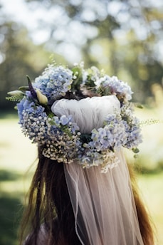 A floral crown made of light blue hydrangeas, white baby's breath, and greenery atop long, flowing hair. The image is taken from behind, with a soft focus background of greenery and dappled sunlight.