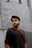 Close-up of a man wearing a charcoal sheiye t-shirt, standing against a gritty urban wall.