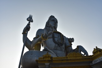 A large statue of a deity sitting with a trident in one hand and the other hand raised in a blessing gesture. The statue is adorned with necklaces and arm bracelets, and features intricate detailing on its base with golden accents. The sky is clear and blue in the background.