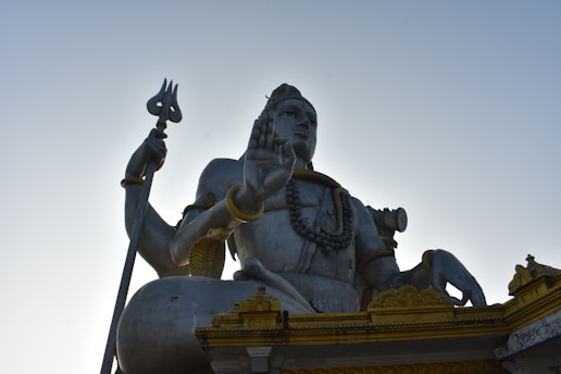 A large statue of a deity sitting with a trident in one hand and the other hand raised in a blessing gesture. The statue is adorned with necklaces and arm bracelets, and features intricate detailing on its base with golden accents. The sky is clear and blue in the background.