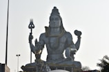 A large statue of a deity sits in a meditative pose. The deity has a calm and serene face, with long hair tied up. There is a snake draped around the neck and multiple necklaces adorn the chest. The deity holds a trident in one hand and another object in the other hand. Smaller statues and various objects are seen at the base of the deity.