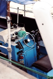 A close-up view of a boat's helm featuring a steering wheel and various controls including radios and gauges. The panel is mostly blue with white accents. There are various marine equipment and a fire extinguisher is visible beneath the console.