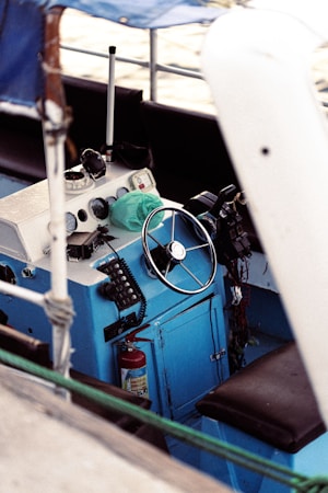 A close-up view of a boat's helm featuring a steering wheel and various controls including radios and gauges. The panel is mostly blue with white accents. There are various marine equipment and a fire extinguisher is visible beneath the console.