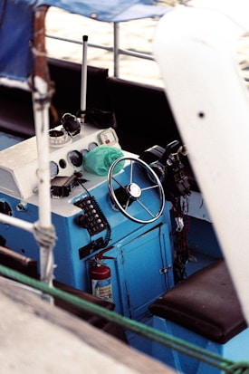A close-up view of a boat's helm featuring a steering wheel and various controls including radios and gauges. The panel is mostly blue with white accents. There are various marine equipment and a fire extinguisher is visible beneath the console.