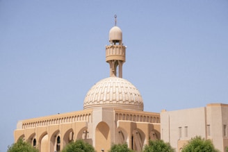 A beige mosque with a large ornate dome and tall minaret set against a clear blue sky. The architecture features arches and intricate designs typical of Islamic structures. Green trees partially frame the building in the foreground.