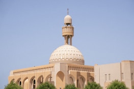 A beige mosque with a large ornate dome and tall minaret set against a clear blue sky. The architecture features arches and intricate designs typical of Islamic structures. Green trees partially frame the building in the foreground.
