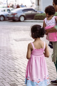 A young girl with curly hair is wearing a pink dress with a pattern of boats along the bottom, standing on a paved sidewalk. Nearby, an adult holds a small child who is dressed in a pink dress with white polka dots. In the background, parked cars are visible, suggesting an urban environment.