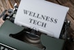 A typewriter with a sheet of paper in the carriage displaying the text 'WELLNESS TECH'. The typewriter is situated on a wooden surface, contributing to a vintage look.